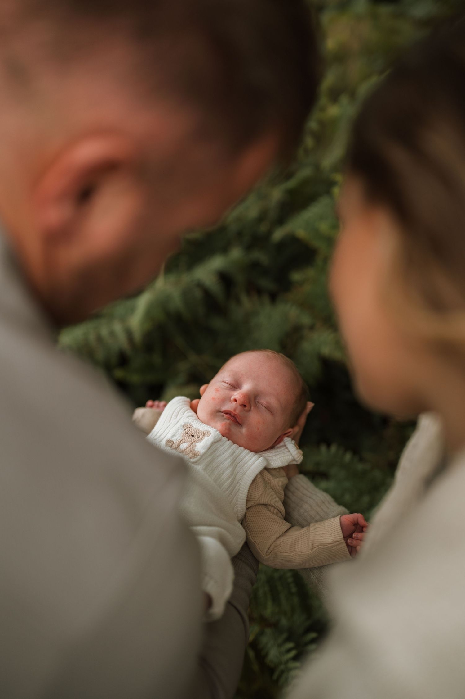 Sleeping newborn baby in a cream outfit seen between two softly blurred parents with green ferns in the background.