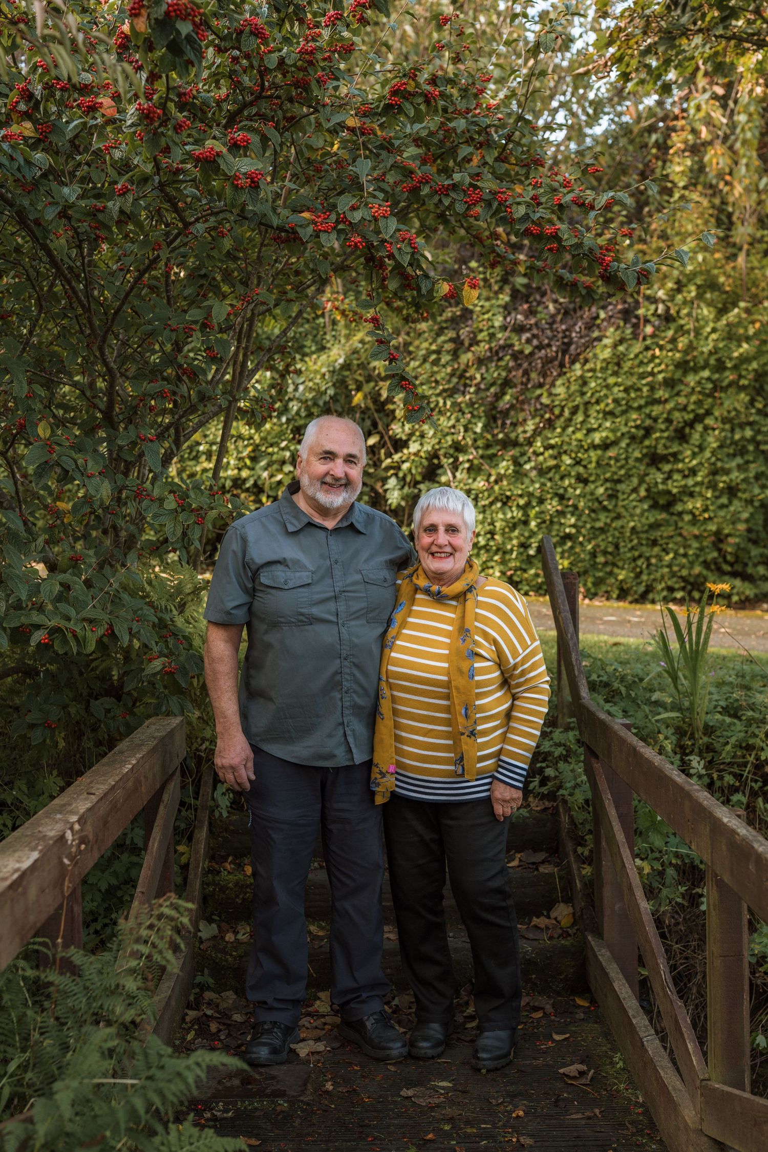 Smiling older couple standing together on a wooden footbridge in a lush autumn garden with red berry-laden trees behind them.