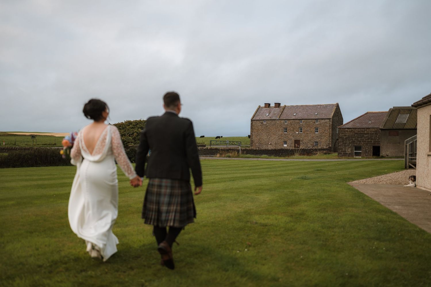 Bride in a white gown and groom in a kilt walk hand in hand across a green lawn toward old stone farm buildings.