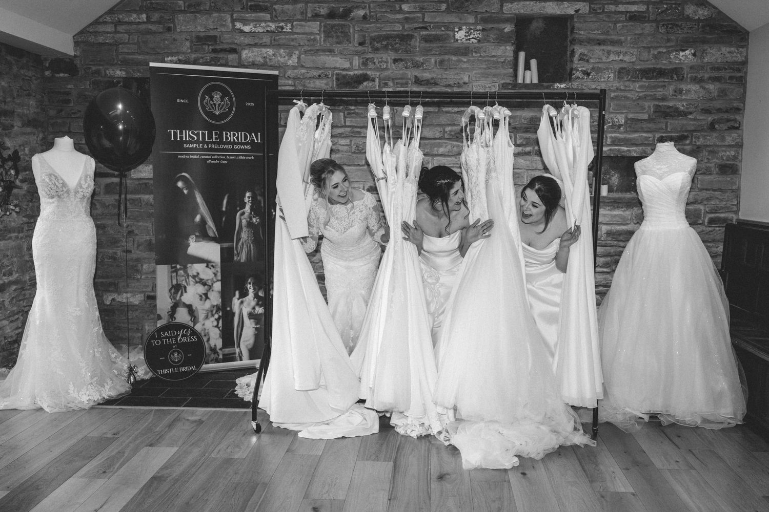 Black and white photo of three women admiring white wedding dresses on a rail in a bridal shop with a Thistle Bridal banner.