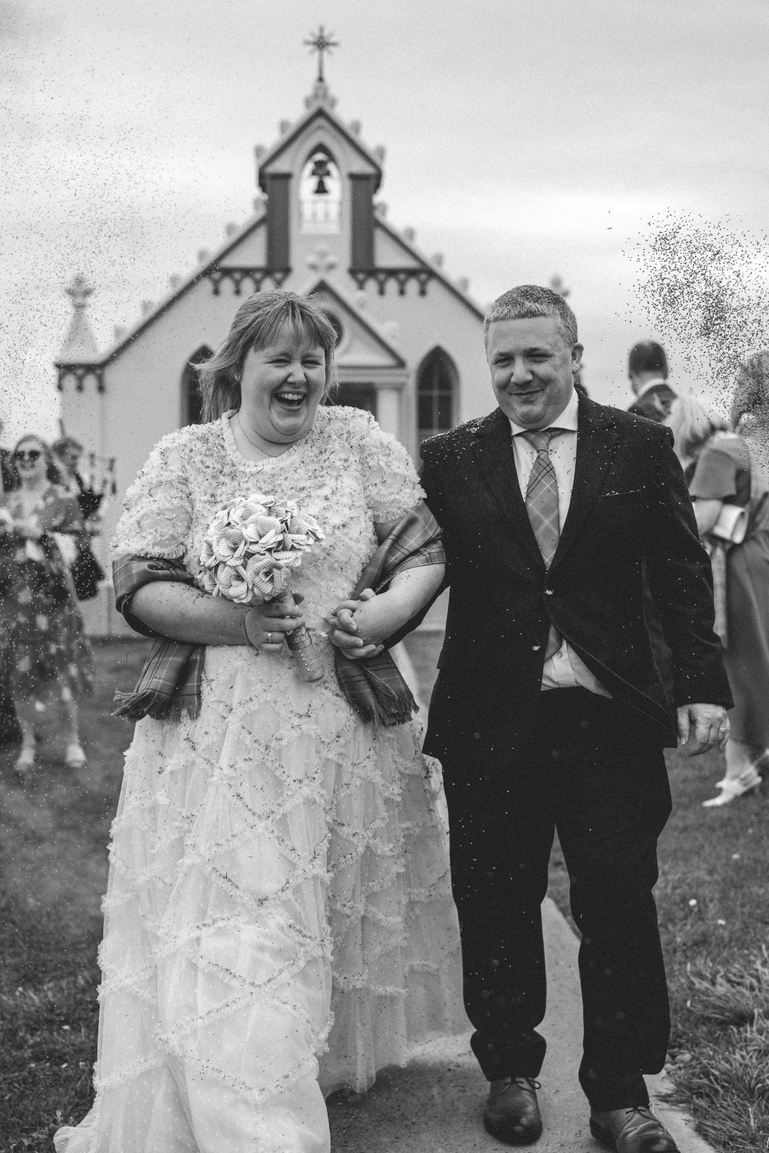 Laughing newlyweds walk arm in arm past a small chapel as confetti falls in black and white.