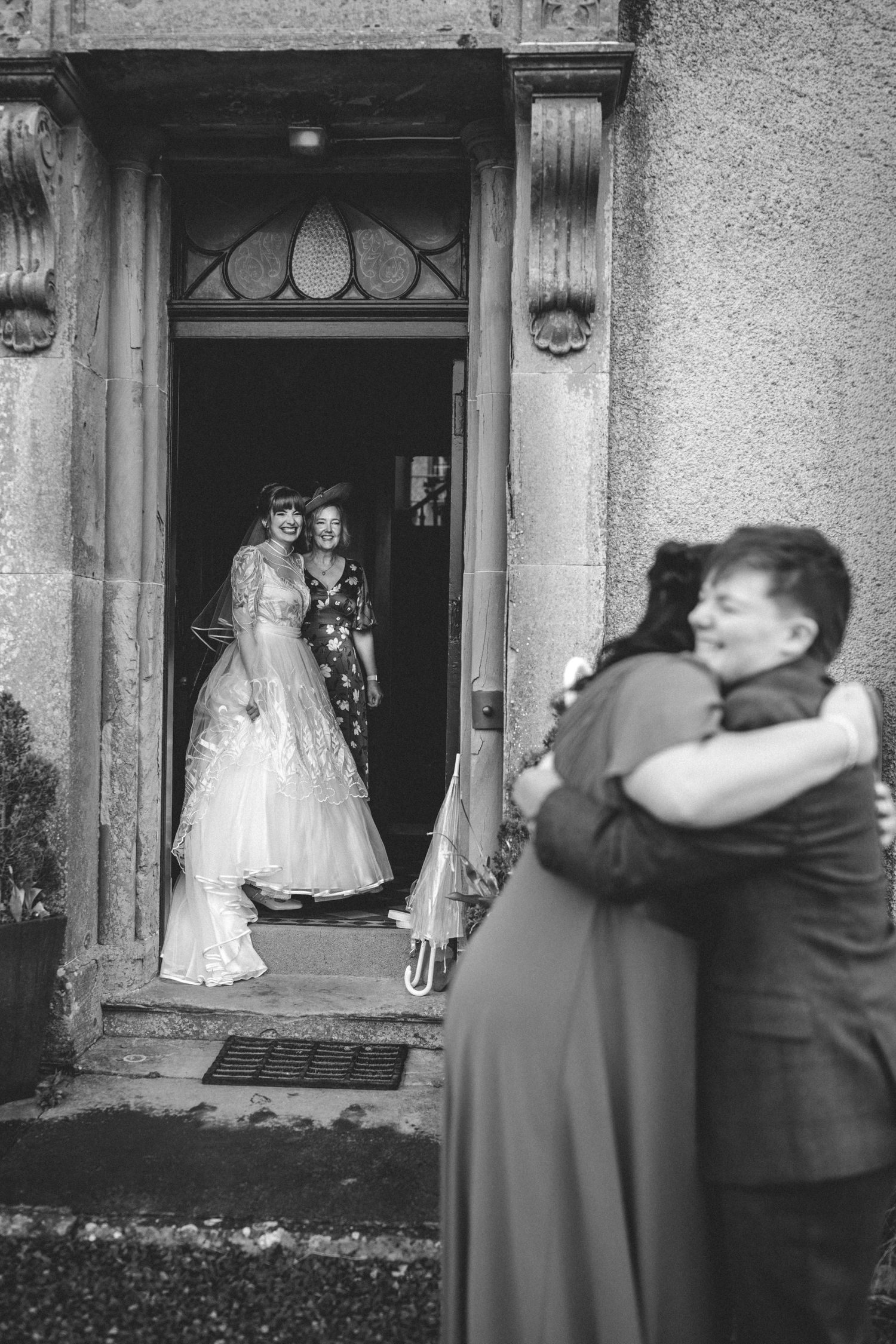 A bride in a lace wedding gown smiles in a stone doorway while two guests share a warm embrace in the foreground.