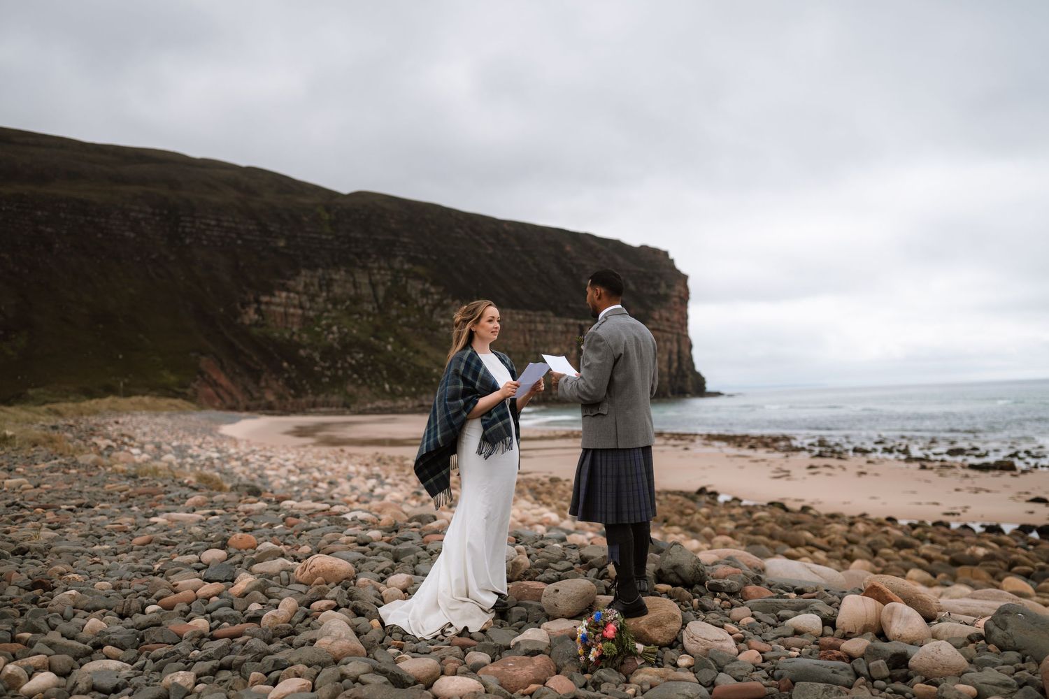Bride in a tartan shawl and groom in a kilt read vows on a pebble beach beneath dramatic coastal cliffs.