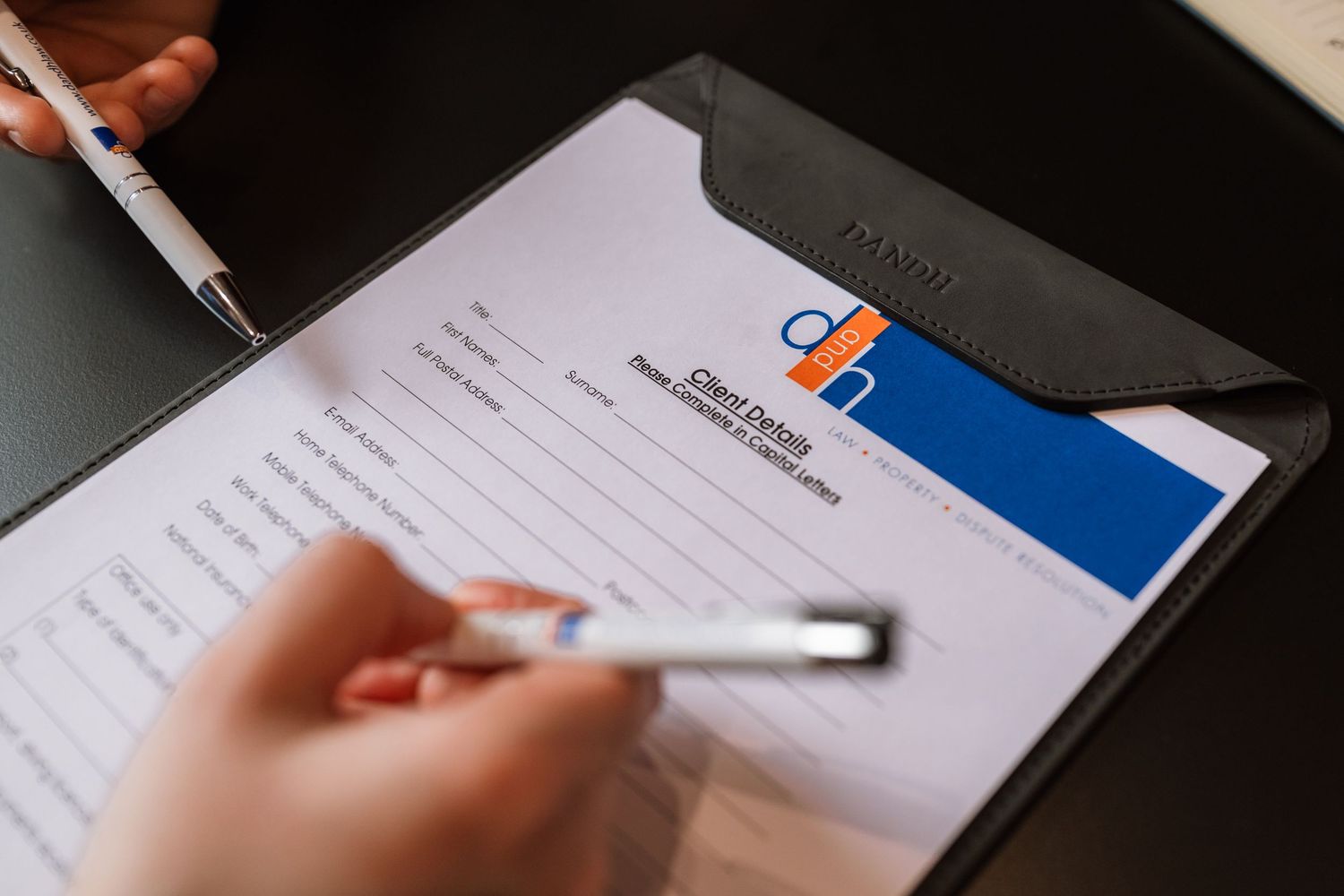 Person filling out a client intake form on a black clipboard with a pen, while another hand holds a second pen nearby.