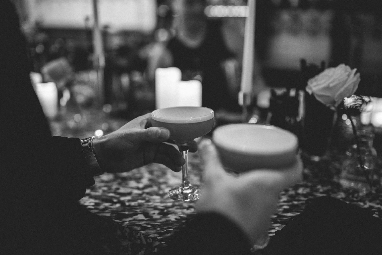 Two hands hold frothy cocktails in coupe glasses at a candlelit bar in a moody black and white photo.