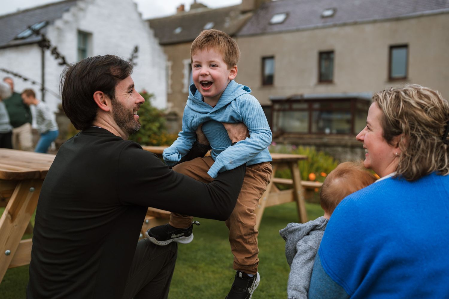 A smiling man lifts a laughing boy in a blue jacket onto his shoulders beside a picnic table as a woman looks on.