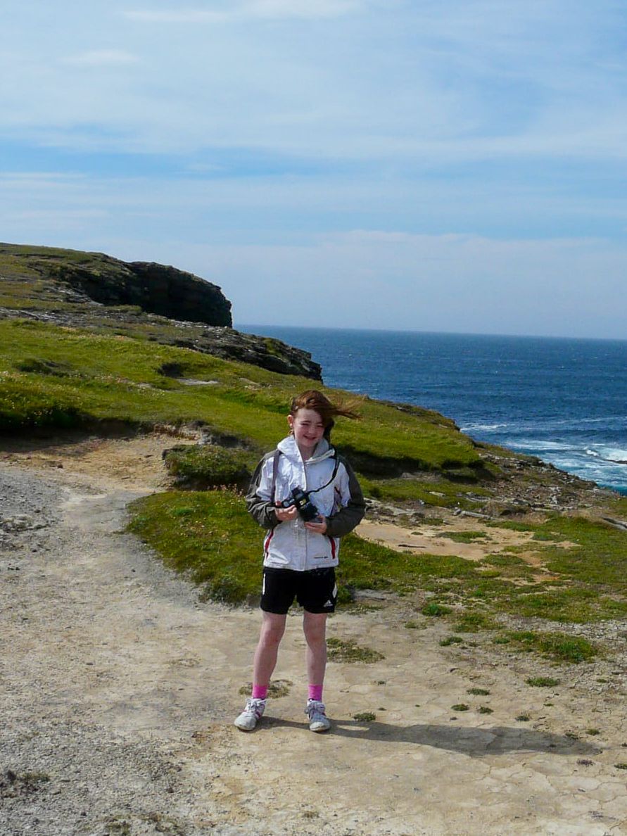 Young girl holding a camera stands on a coastal cliff path with green grass and blue ocean in the background.