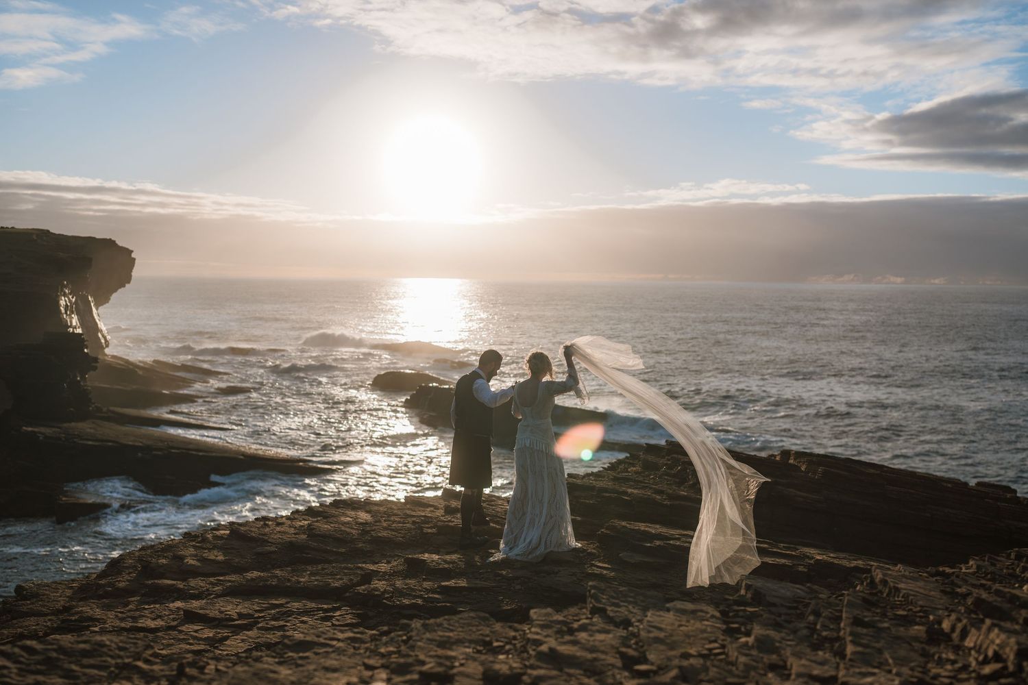 Bride and groom stand on coastal rocks at golden hour as the sun sets over the sea and the bride's veil blows in the wind.