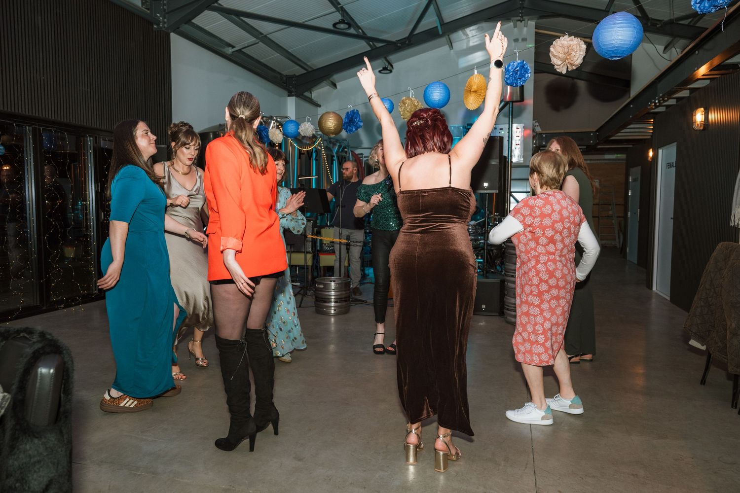 Guests dance at a festive party with blue and gold balloons, including a woman in a velvet brown dress raising her arms.