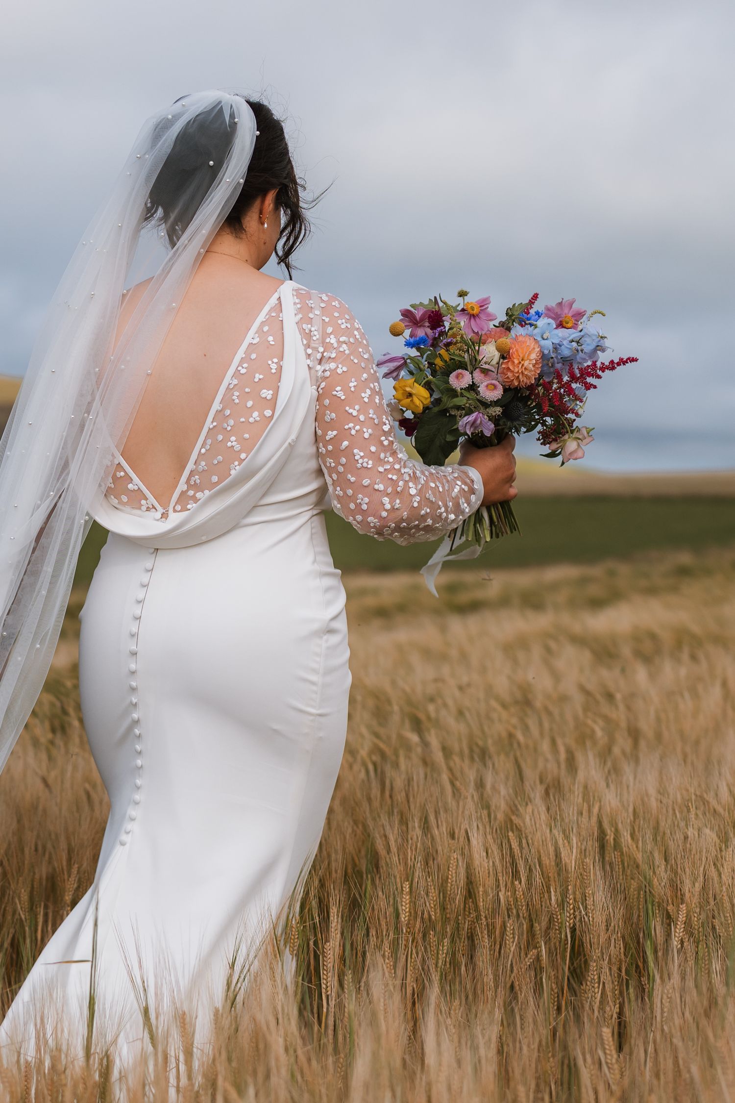 Bride in a lace-sleeved gown and long veil holds a colorful bouquet while standing in a golden barley field.