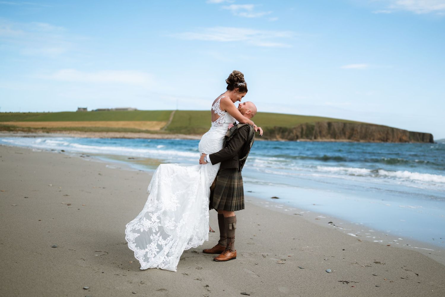 Groom in a kilt lifts his bride in a flowing lace dress on a sandy beach beside turquoise waves and green coastal hills.