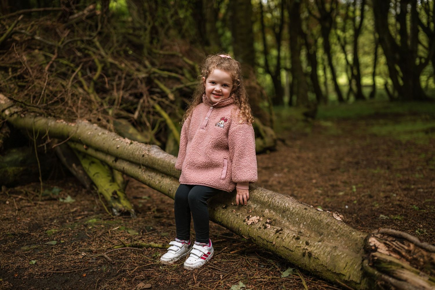 Smiling little girl in a pink fleece jacket sits on a fallen log in the woods.