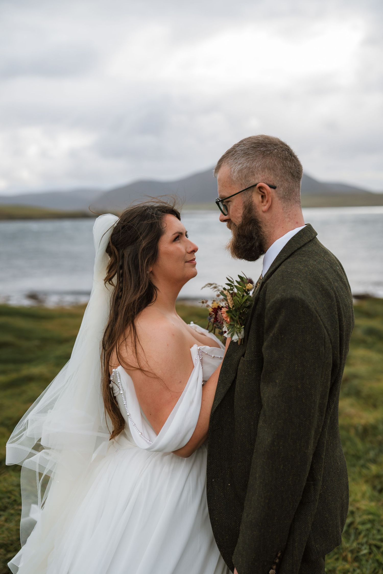 Bride and groom in tweed jacket gaze at each other beside a loch with misty hills behind, her bouquet and veil softly blowing.
