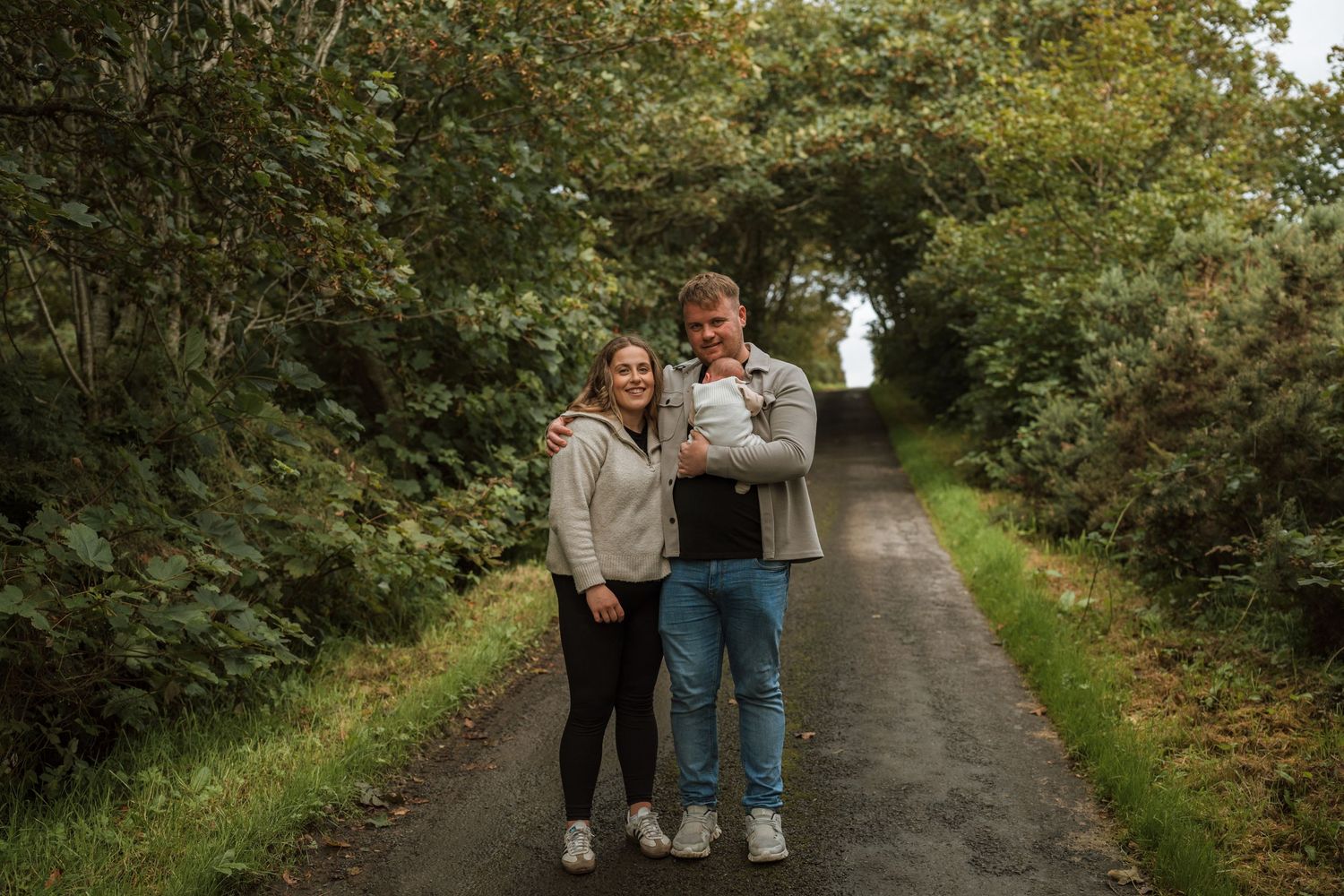New parents standing on a tree-lined country path holding their newborn baby during an outdoor family photo session.