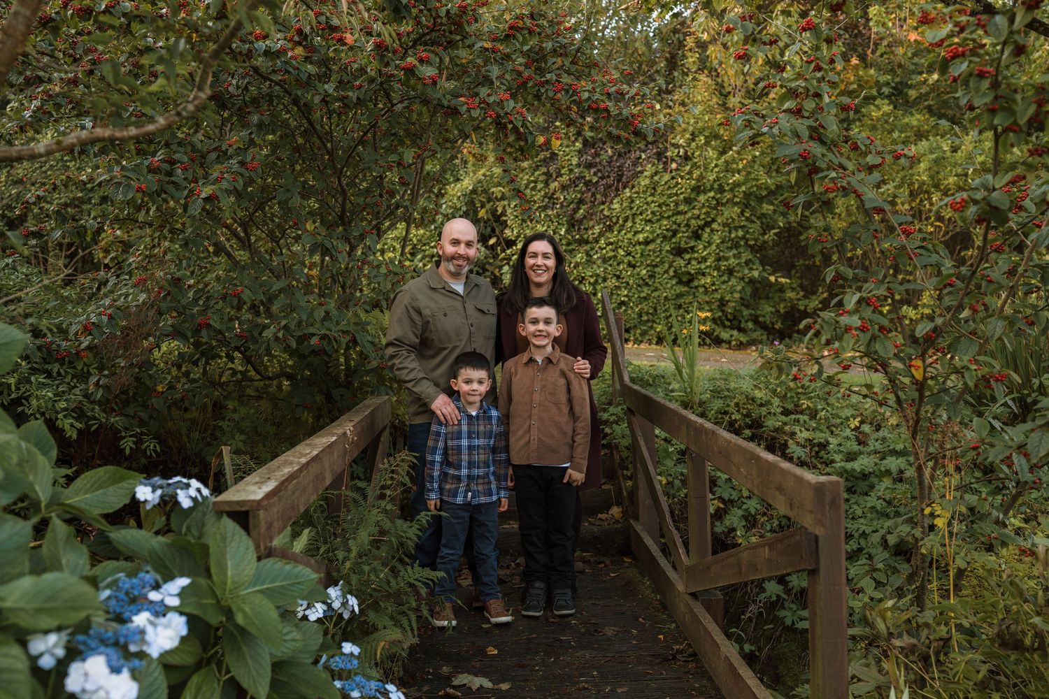 Family of four poses together on a wooden footbridge surrounded by lush green foliage and blue hydrangeas in a peaceful garden setting.