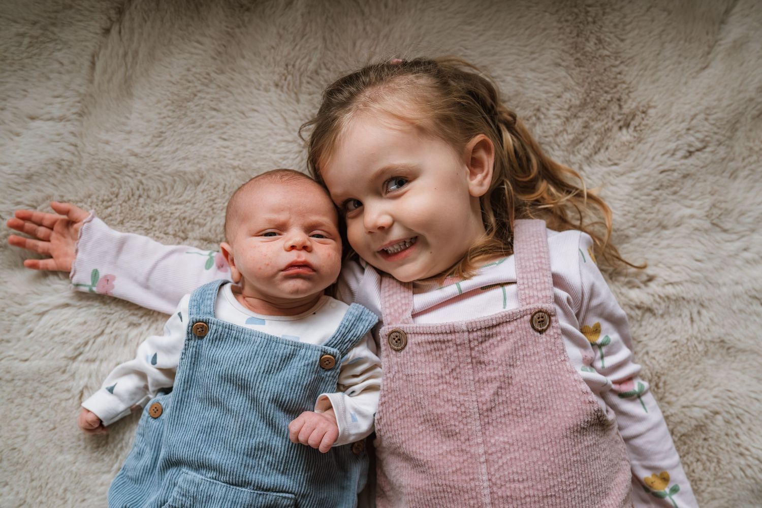Smiling toddler girl in a pink pinafore lies on a soft cream rug beside her sleeping newborn sibling in a blue denim overall.