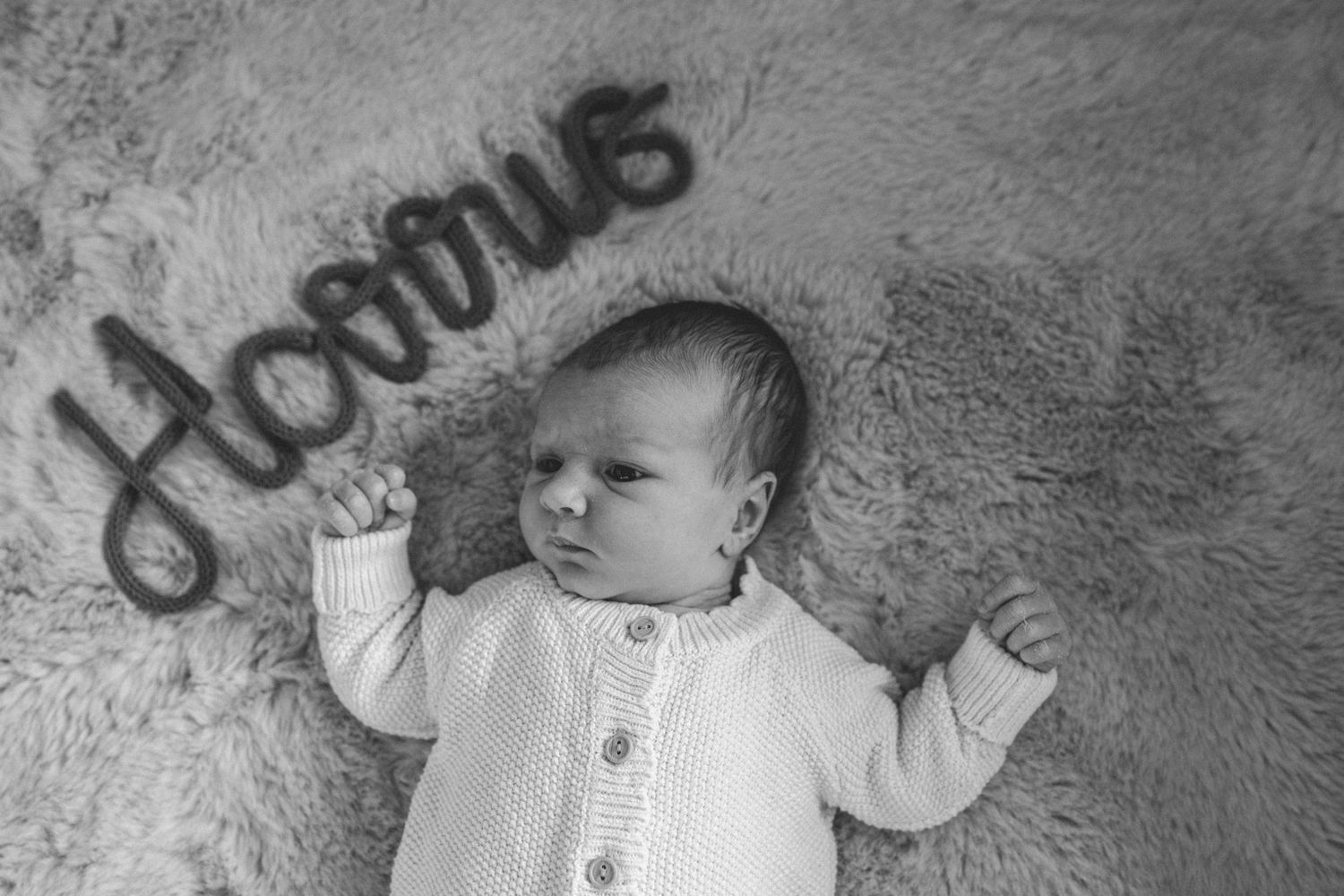 Black and white photo of a newborn baby in a knit cardigan lying on a fluffy rug beside decorative wire lettering spelling a name.