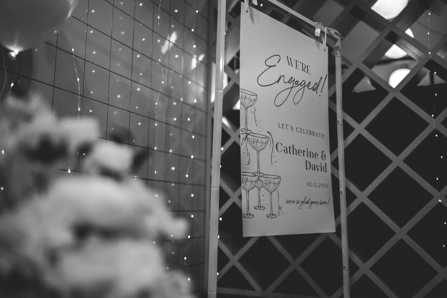 Black and white photo of a We're Engaged sign for Catherine & David with champagne glasses, hung on a lattice near string lights.