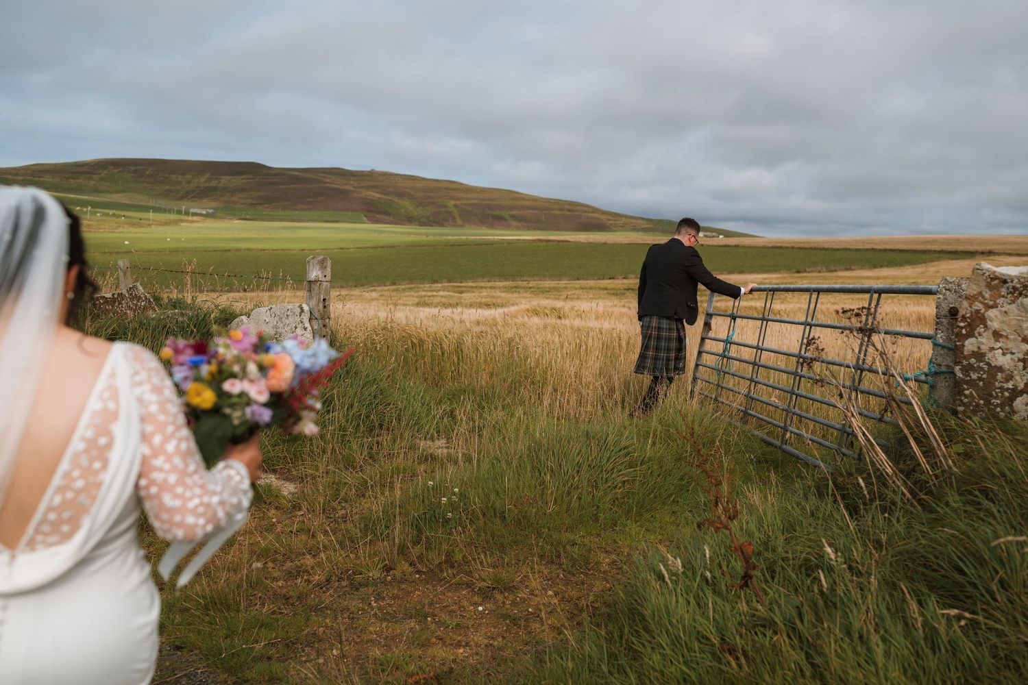Bride holding a colorful bouquet watches a groom in a kilt open a metal farm gate in a Scottish countryside field.
