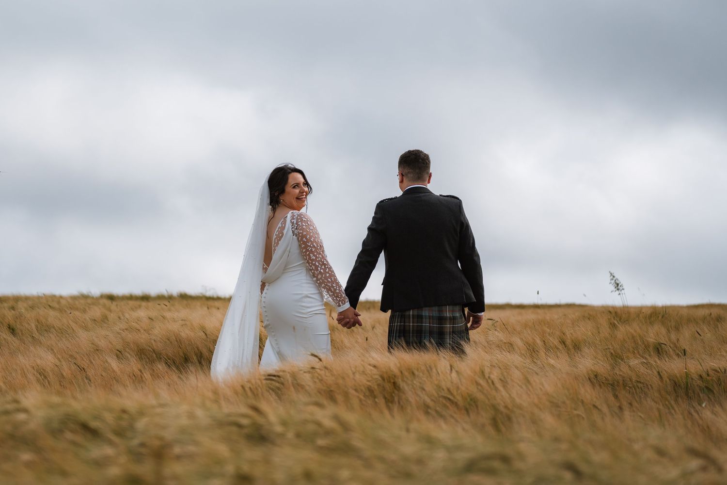 A bride in a veil and a groom in a kilt hold hands and walk through golden grass under a cloudy sky.