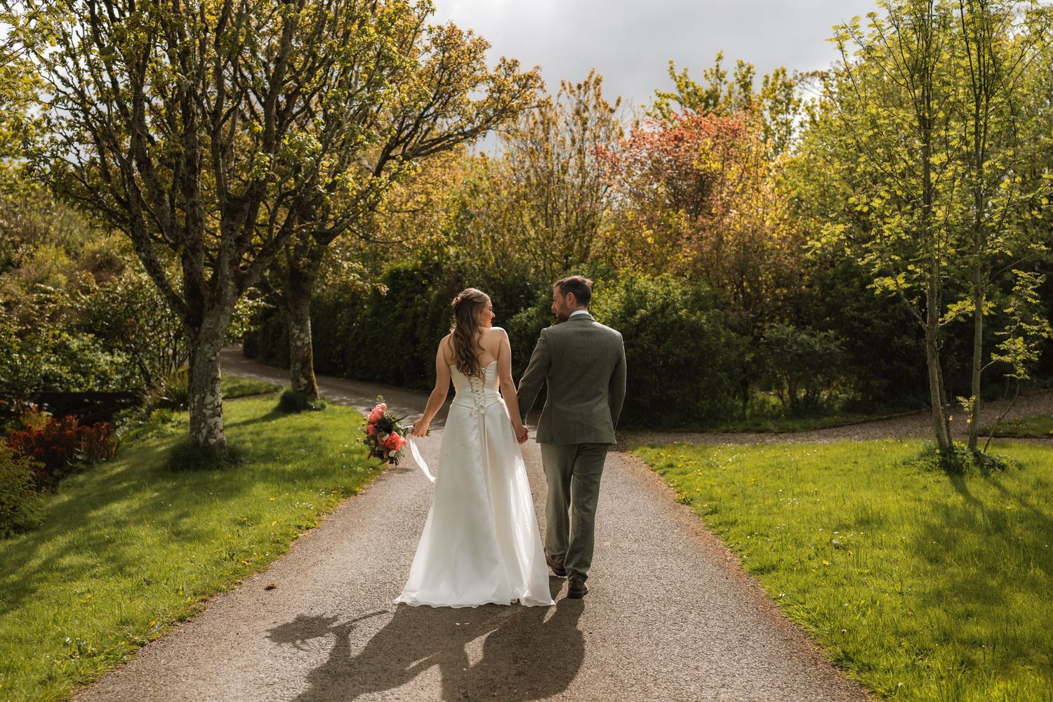 A bride in a white gown holds a bouquet and the groom's hand as they walk down a sunlit garden path surrounded by trees.