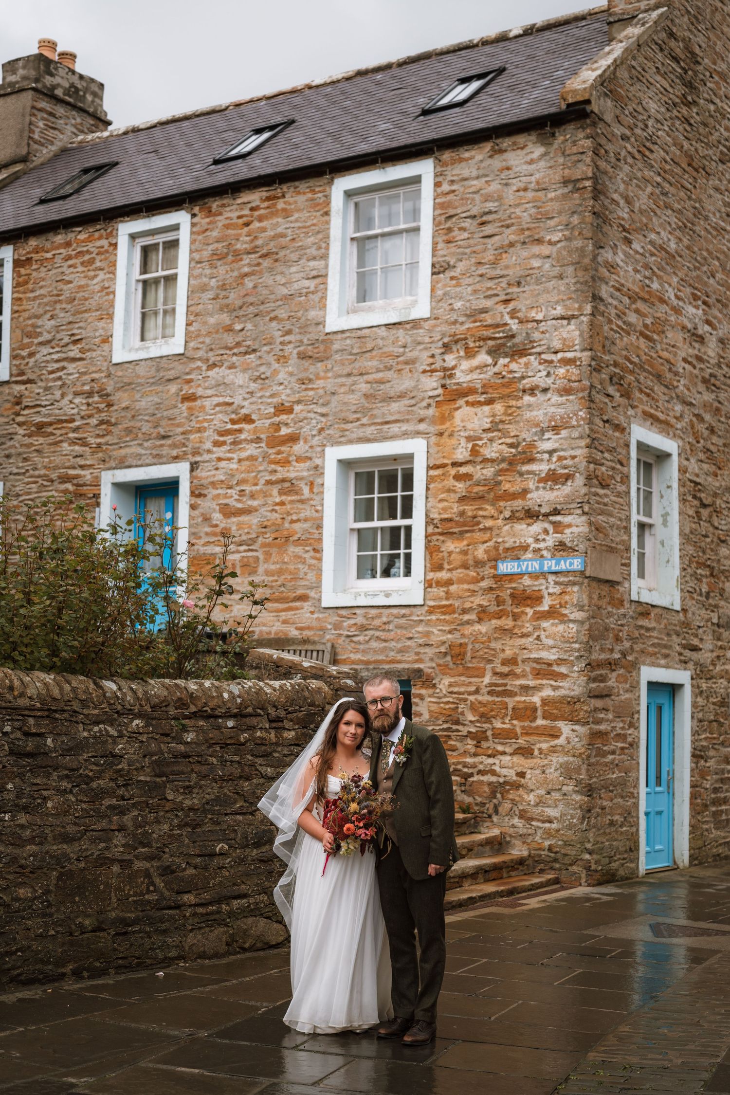 Bride in a long veil and groom in a tweed suit pose holding a bouquet outside a stone house with blue doors on a wet street.