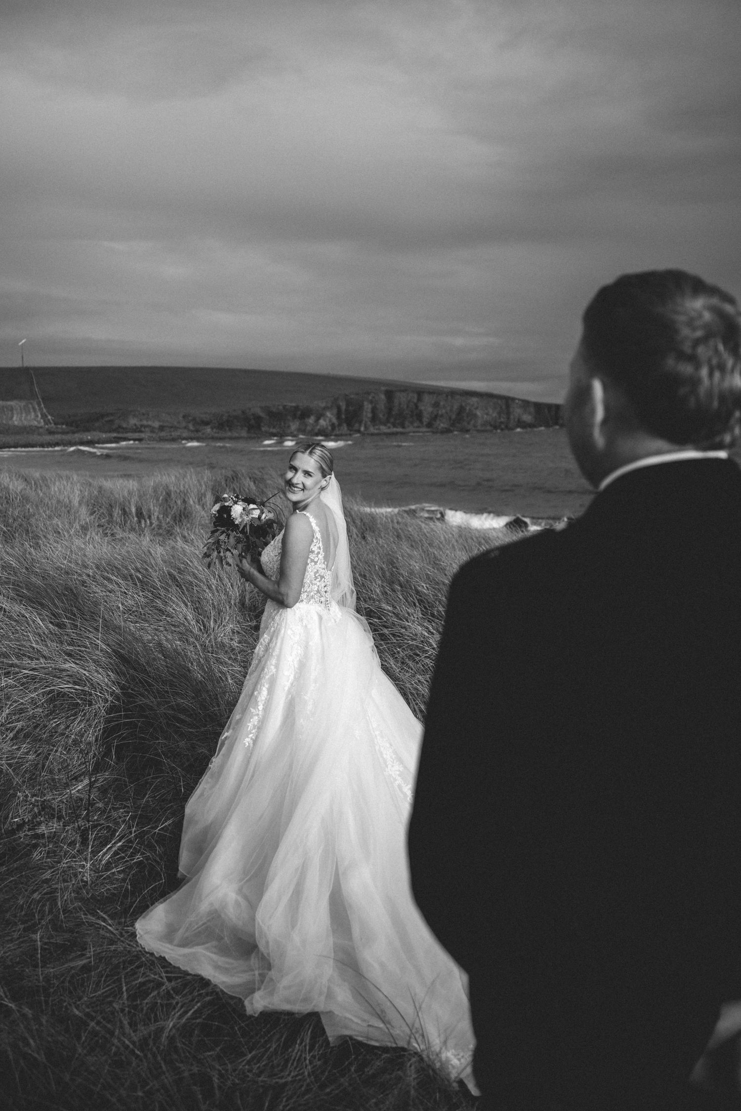 Bride in a flowing tulle gown holding a bouquet smiles over her shoulder at the groom in dune grass overlooking a moody coastal cliff.