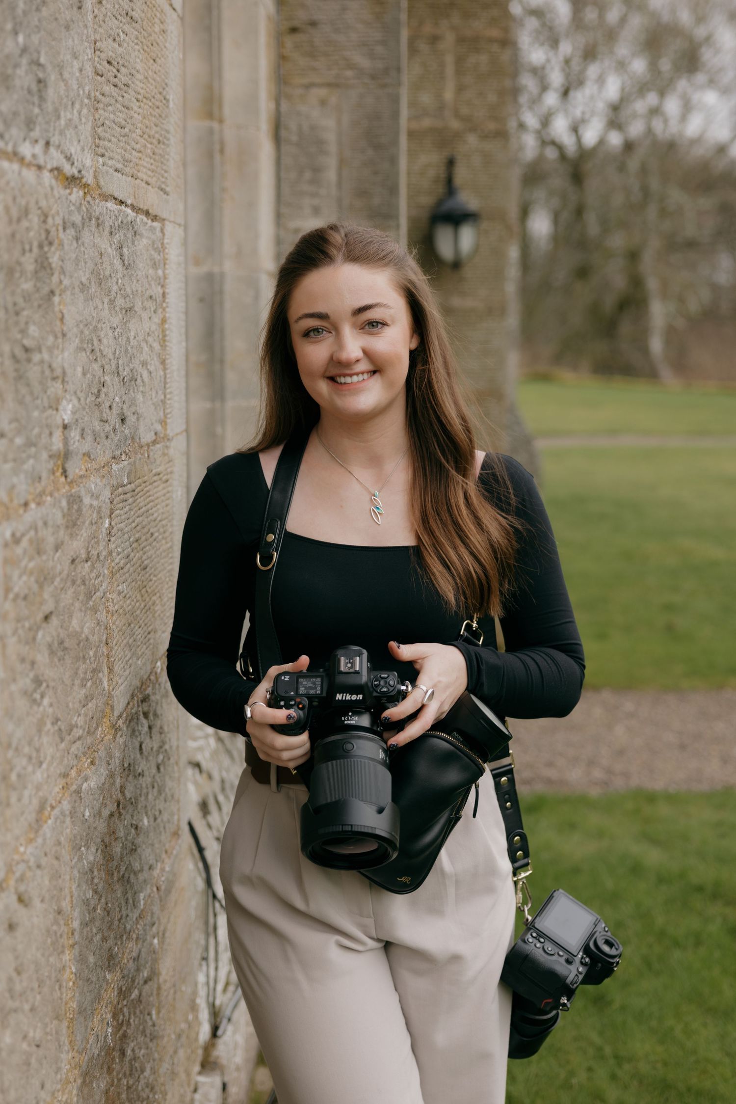 Smiling woman with long brown hair leaning against a stone wall holding a Nikon camera, with a second camera at her side.