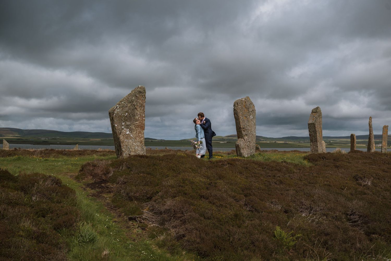Wedding couple embrace among ancient standing stones on a heather-covered hill beneath a moody, cloudy sky.