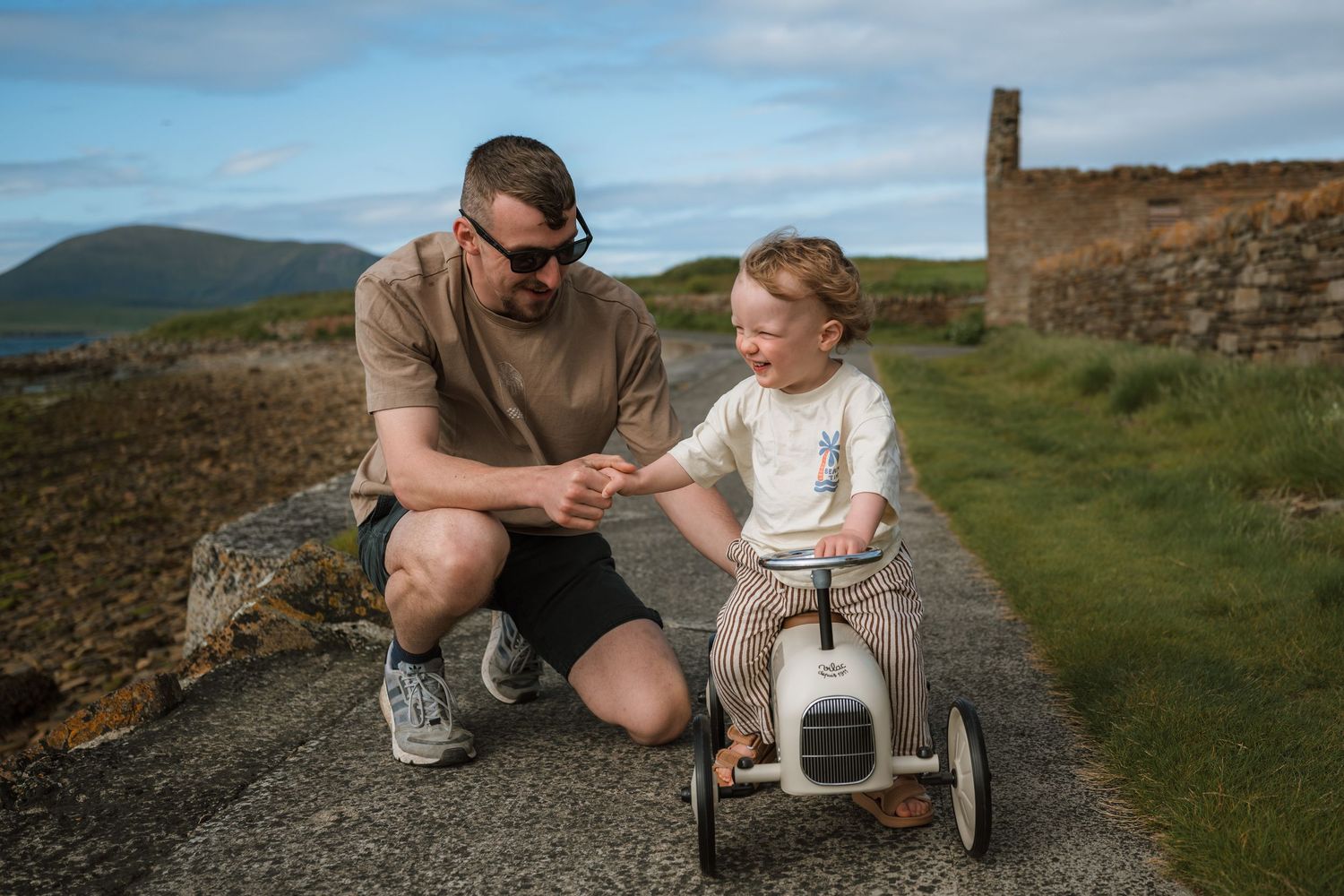 A smiling man crouches to hold the hand of a toddler riding a small vintage-style balance bike along a coastal path.