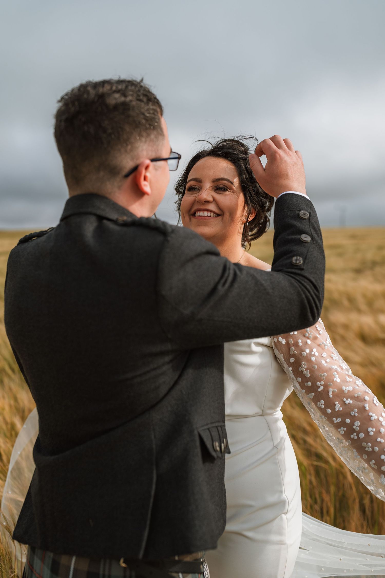 Smiling bride in a white gown with sheer beaded sleeves as the groom in a tweed jacket and kilt adjusts her hair in a golden field.