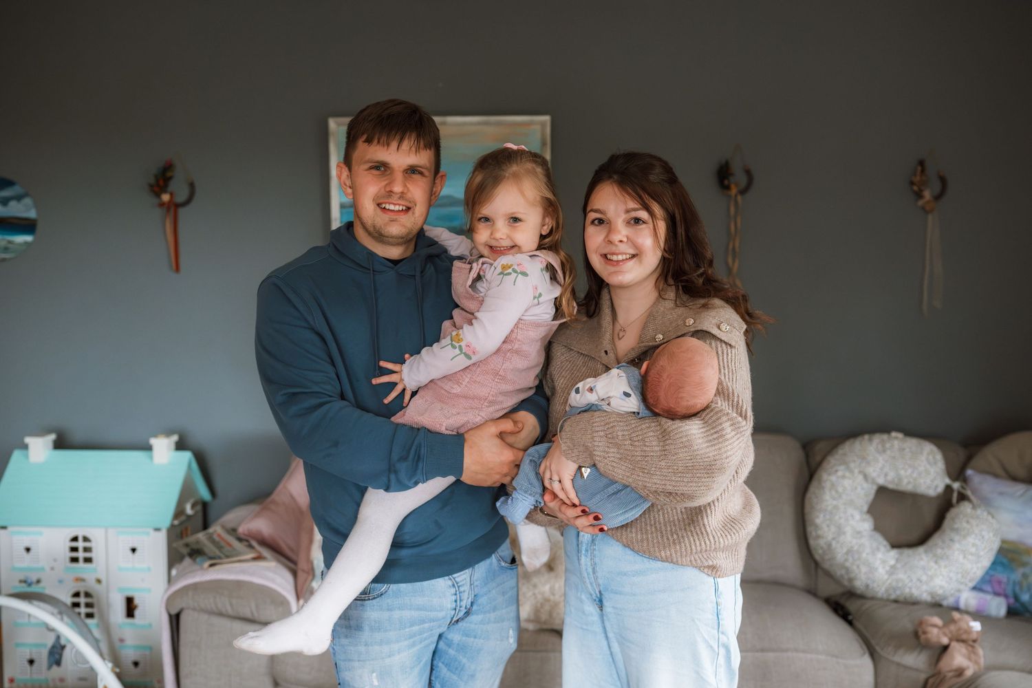 Smiling young family of four in a cozy living room, with a father holding a toddler and a mother cradling a newborn baby.