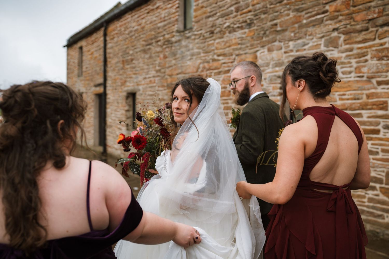 Bridesmaids in dark dresses adjust a bride's long veil beside a rustic stone building while a bearded groom looks on.