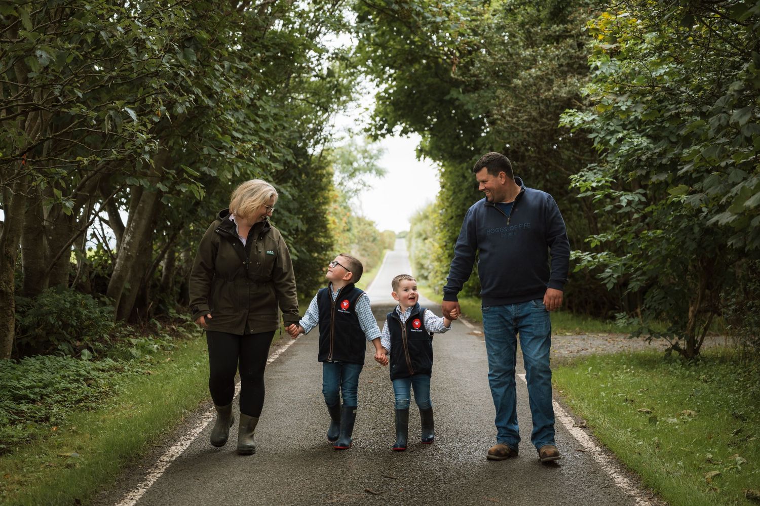 A man, a woman and two children hold hands while walking down a tree-lined country path in autumn.