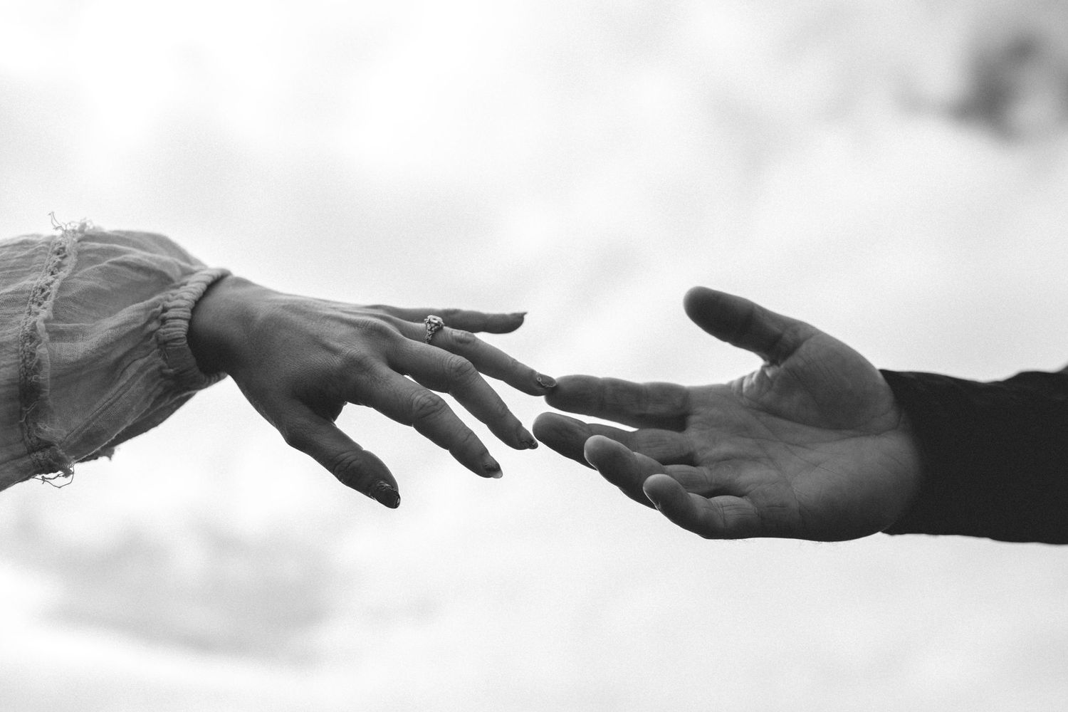 Black and white image of two hands reaching toward each other against a soft cloudy sky.