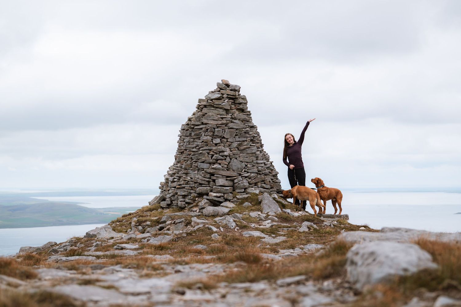 Woman raises her arm in celebration beside two golden dogs at a stone cairn on a hilltop above a misty coastline.