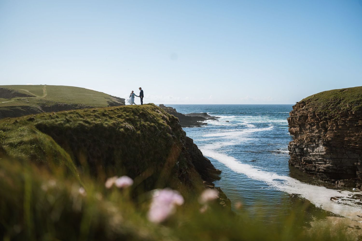 A couple in wedding attire stand on a grassy clifftop overlooking turquoise waves, with pink wildflowers in the foreground.