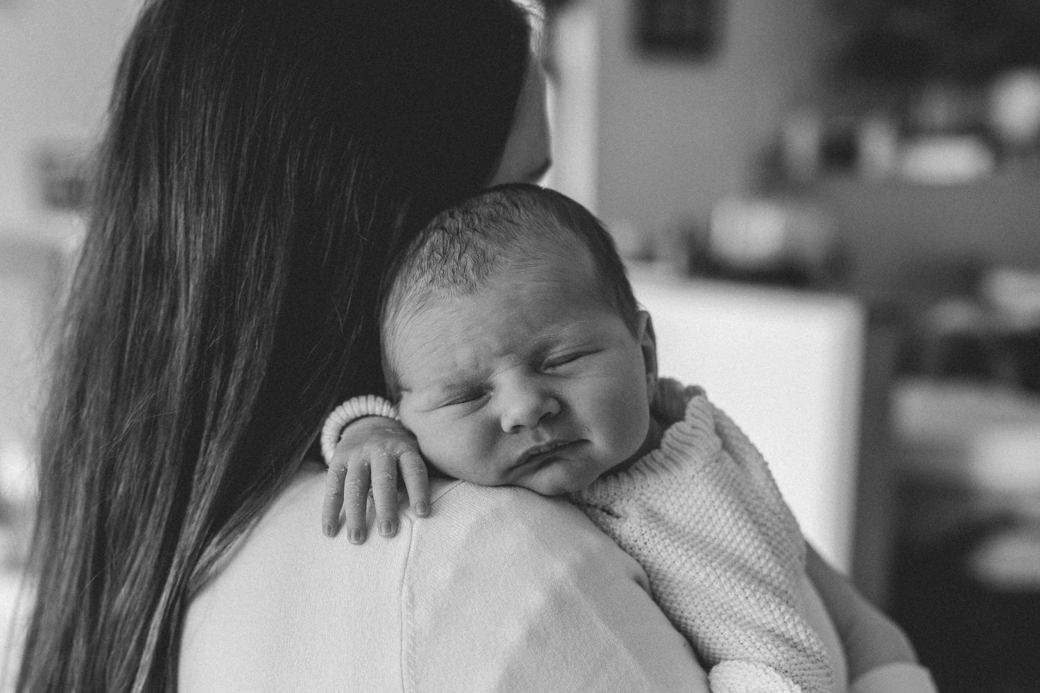 Black and white photo of a sleeping newborn baby resting on the shoulder of a long-haired mother in a cozy home setting.