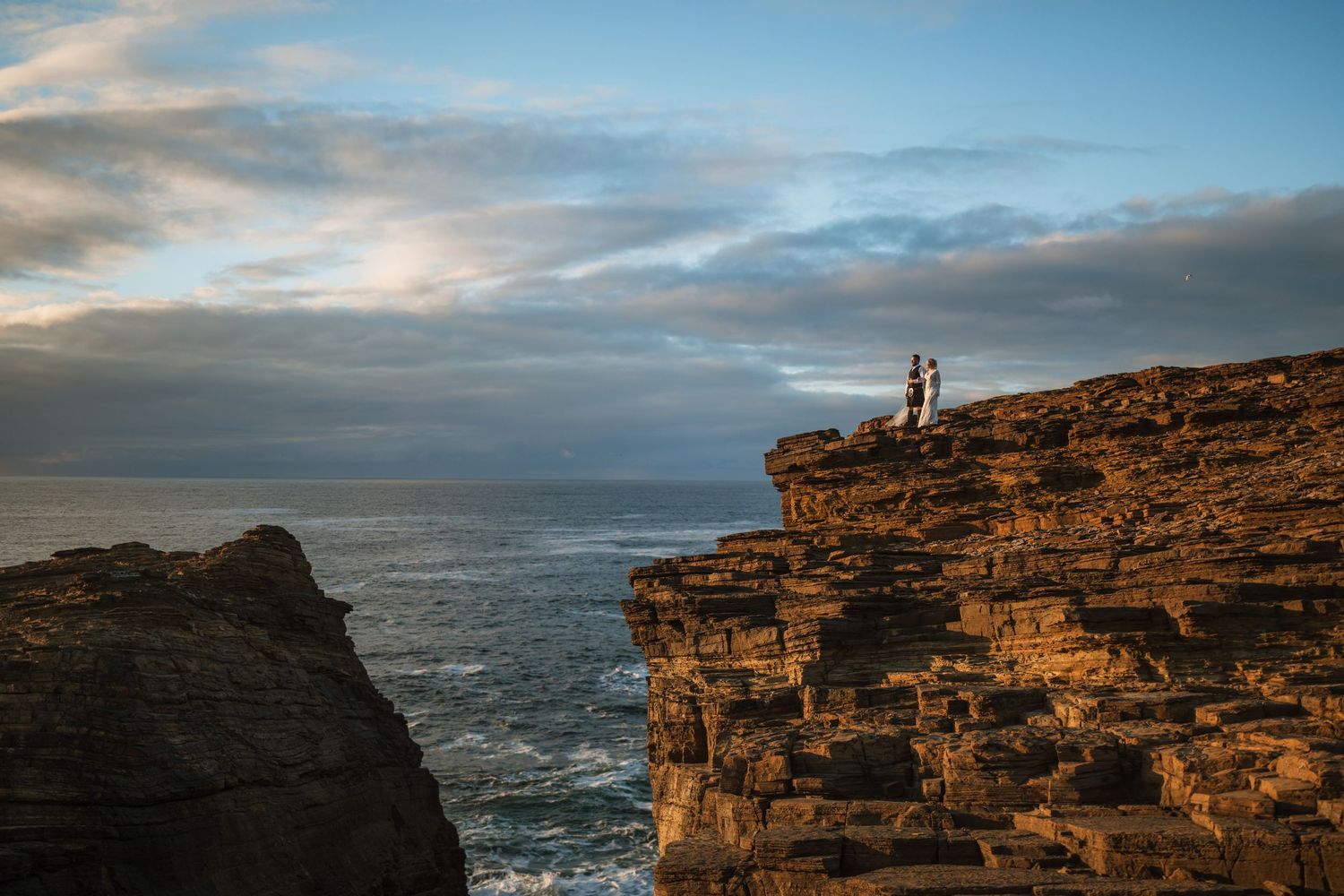 Couple standing on a rugged sea cliff edge at golden hour overlooking the ocean under a dramatic evening sky.