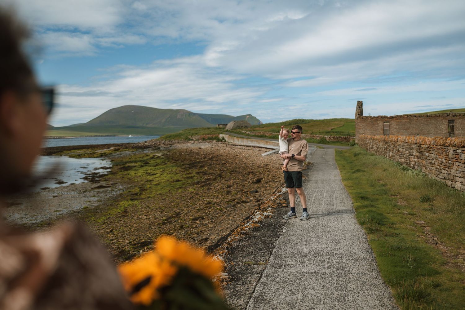 A couple walks along a coastal path beside an old stone wall with green hills and sea in the background, yellow flowers in foreground.