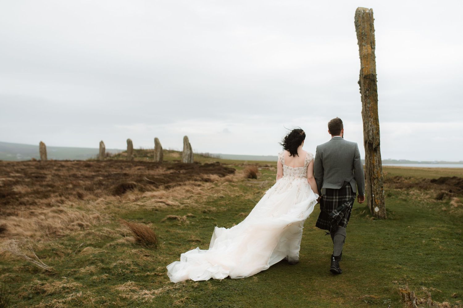 Bride in a flowing white dress and groom in a kilt walk toward ancient standing stones on a grassy Scottish moor.