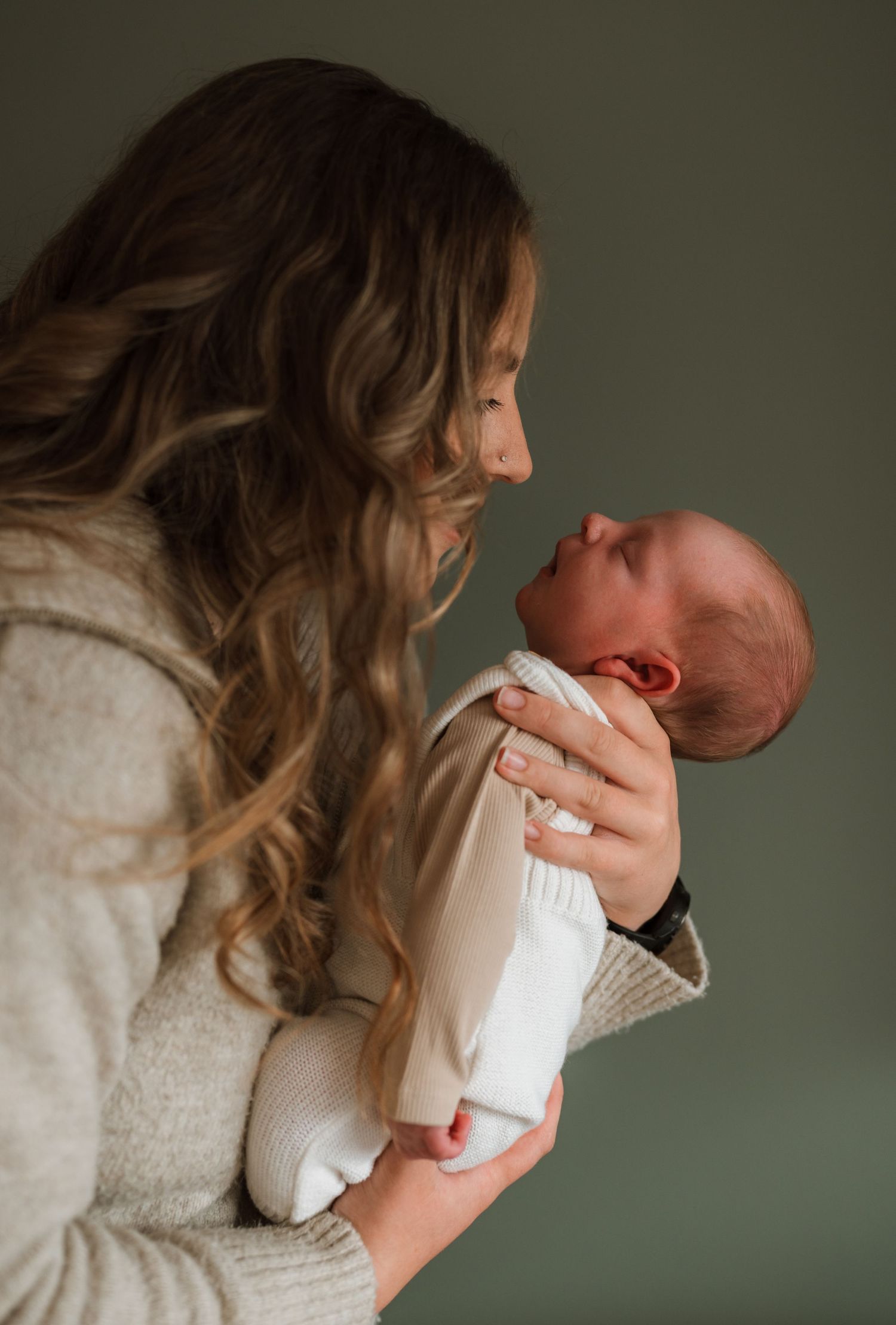 Mother with long wavy hair gently holding her newborn baby up to face her in a soft, moody newborn portrait.