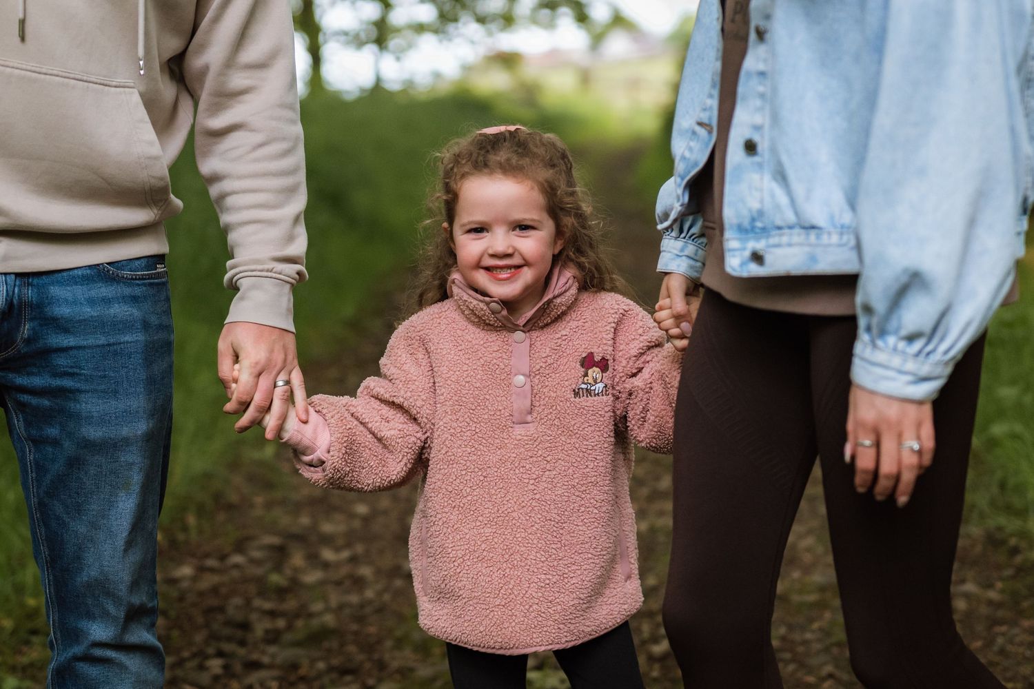 Smiling little girl in a pink fleece holds hands with two parents while walking along a woodland path.