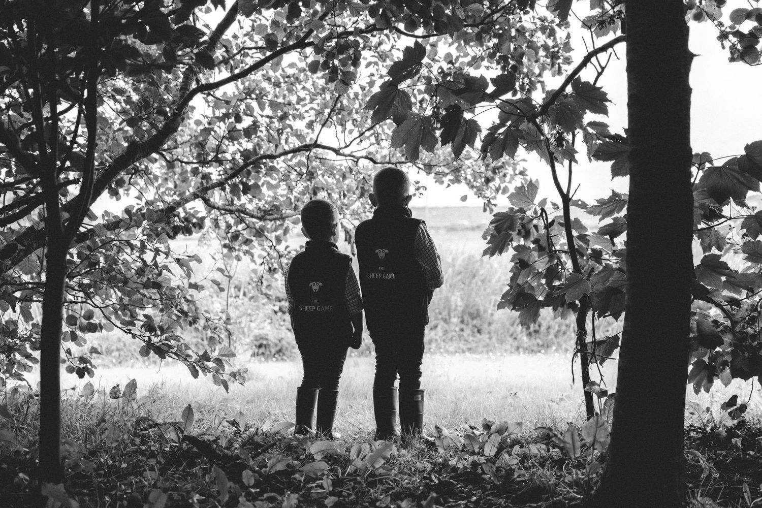Two children wearing backpacks stand among trees looking out over an open field in a black and white woodland photograph.