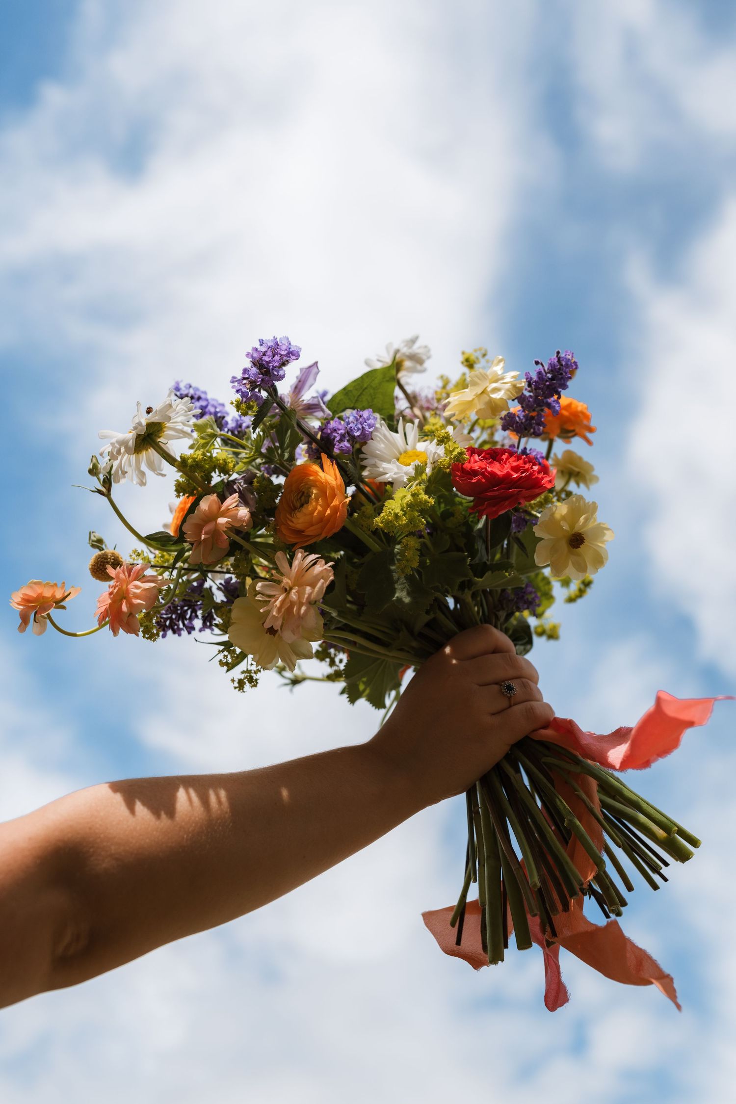 A hand holds up a colorful wildflower bouquet tied with coral ribbons against a bright blue sky with soft white clouds.