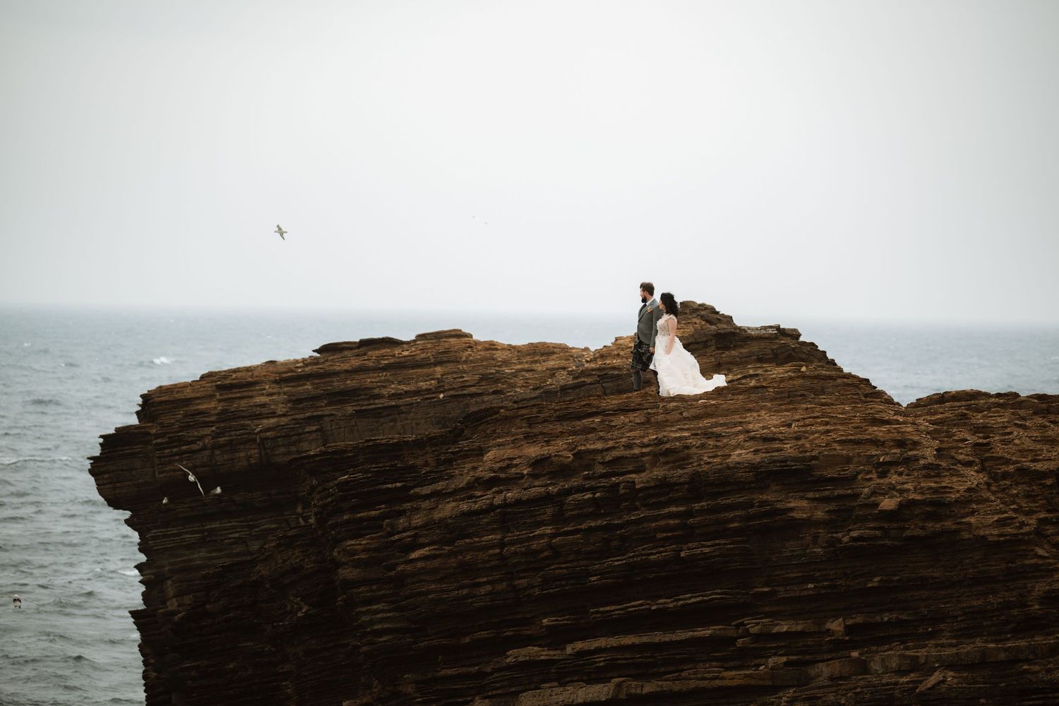 Distant bride and groom stand on top of a rugged sea cliff overlooking the ocean on a hazy day.