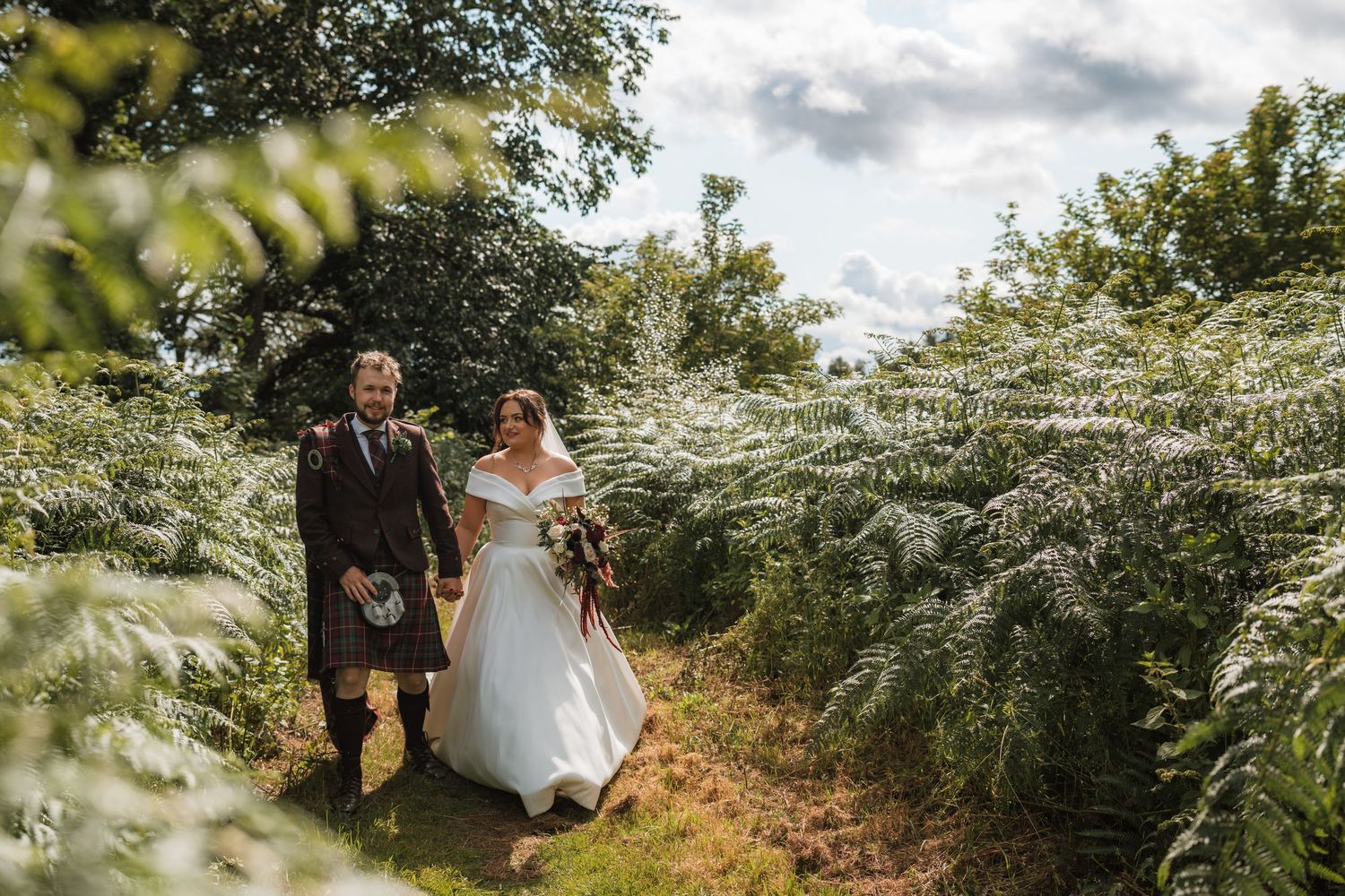 Groom in a kilt and bride in a white dress holding a bouquet walk hand in hand along a grassy path lined with ferns.