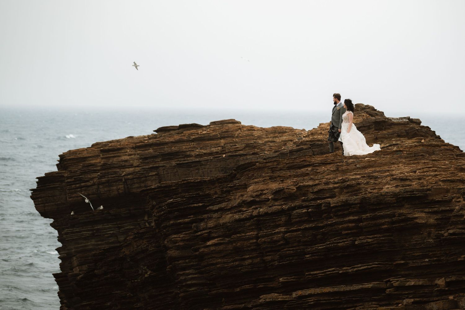 Bride in a white gown and groom standing on a rugged coastal cliff overlooking the misty sea as a seagull flies past.