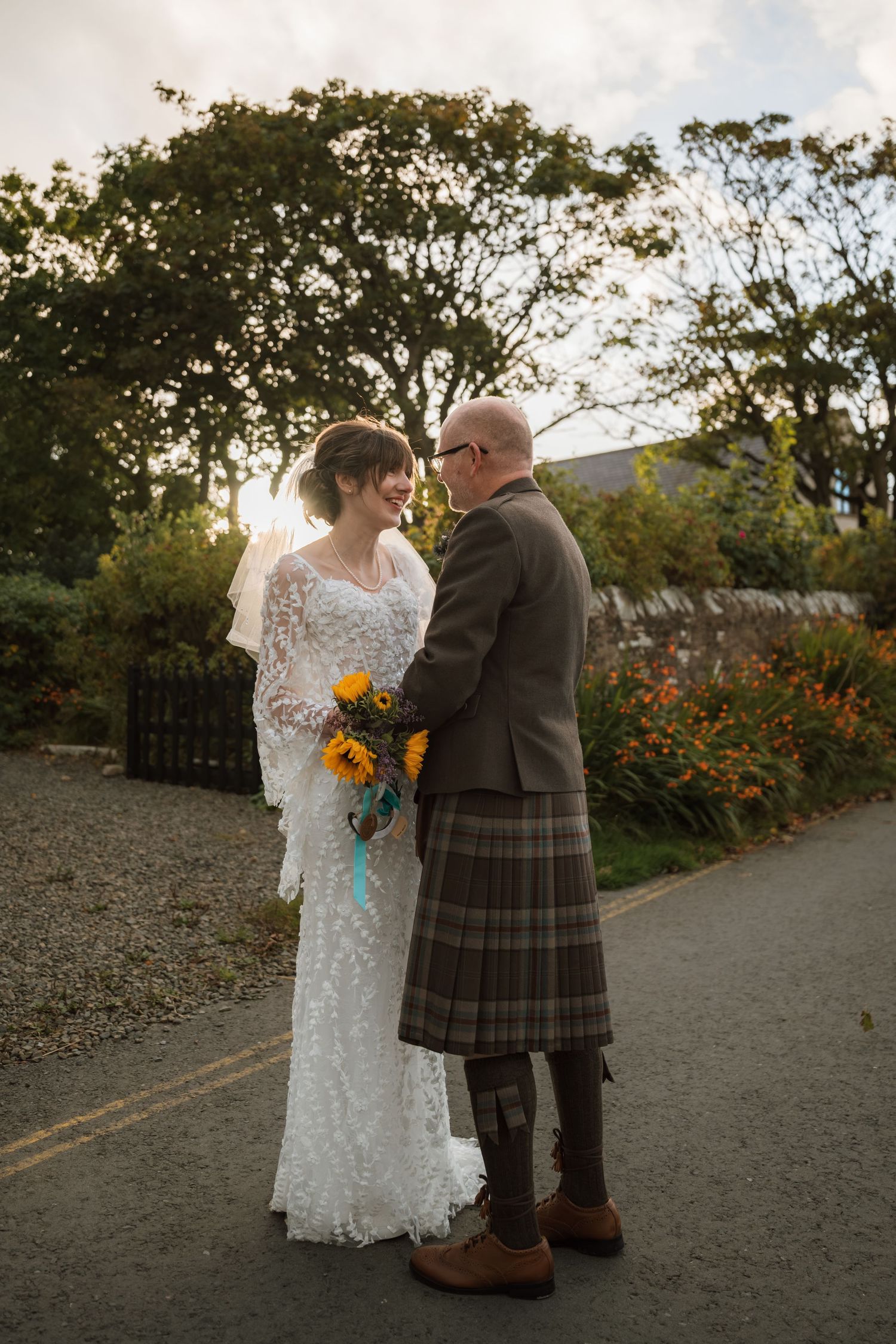 Bride holding a sunflower bouquet faces her groom in a tartan kilt on a quiet road at sunset.