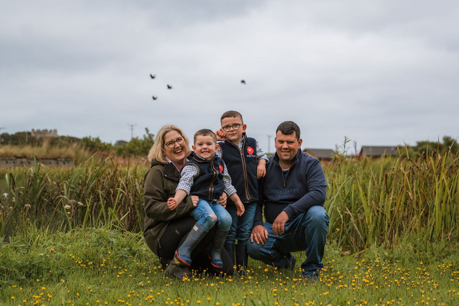 A family of four crouches together in a grassy meadow with wildflowers while birds fly overhead on a cloudy day.